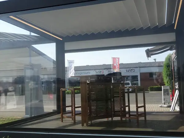 Moderne Terrassenüberdachung mit Glaswänden und Holz-Gartenmöbeln bei Tageslicht.