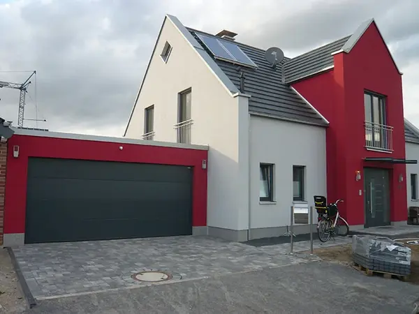 Modernes Einfamilienhaus mit Garage, Solaranlage und roter Fassade auf gepflastertem Hof.